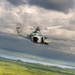 A military helicopter, appearing to be a Bell UH-1Y Venom (Super Huey), is captured in flight over a lush green forested landscape with a body of water visible in the background. The aircraft is painted in a light gray military scheme and has its side door open. The image is taken from another airborne platform, suggesting an air-to-air photography mission.