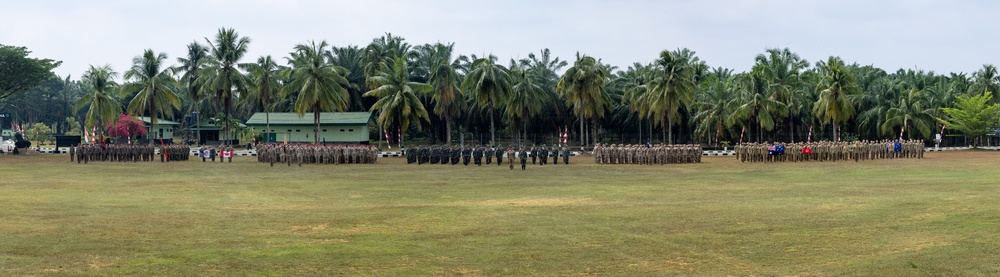 A wide panoramic view of a military parade or formation ceremony taking place on a large grass field. Multiple units of uniformed personnel are arranged in organized rectangular formations across the parade ground. The setting appears to be a tropical military base, evidenced by the palm trees and lush vegetation in the background.