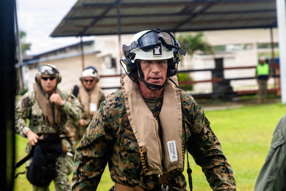 The image shows military personnel in full tactical gear moving purposefully across a grassy outdoor area near a building. The lead figure wears MARPAT woodland camouflage, a white flight helmet with dark visor, communications headset, microphone, and an inflatable life preserver vest. Additional personnel in similar gear follow behind, suggesting a coordinated military operation or exercise.