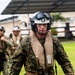 The image shows military personnel in full tactical gear moving purposefully across a grassy outdoor area near a building. The lead figure wears MARPAT woodland camouflage, a white flight helmet with dark visor, communications headset, microphone, and an inflatable life preserver vest. Additional personnel in similar gear follow behind, suggesting a coordinated military operation or exercise.