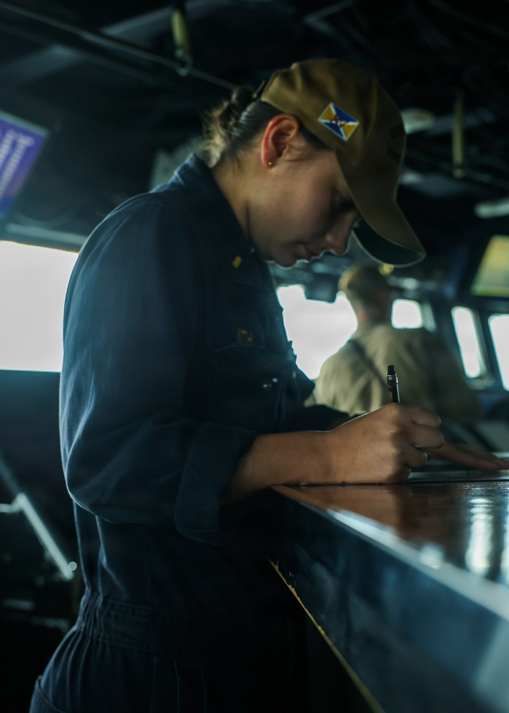A naval personnel member in dark blue uniform is writing or recording data on a surface inside what appears to be a ship's bridge or combat information center. They are wearing a tan/khaki ball cap with a blue and gold insignia patch. A second person in a tan uniform is visible in the background near large windows.