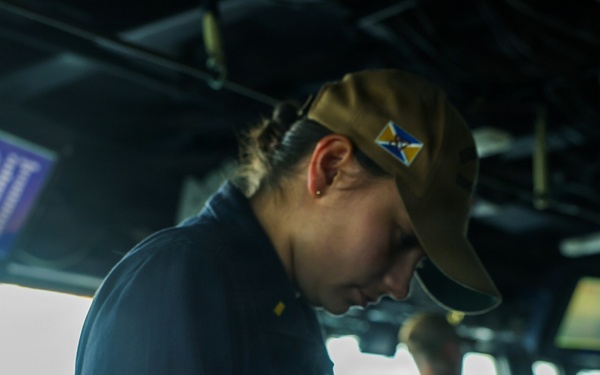 A naval personnel member in dark blue uniform is writing or recording data on a surface inside what appears to be a ship's bridge or combat information center. They are wearing a tan/khaki ball cap with a blue and gold insignia patch. A second person in a tan uniform is visible in the background near large windows.