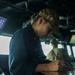 A naval personnel member in dark blue uniform is writing or recording data on a surface inside what appears to be a ship's bridge or combat information center. They are wearing a tan/khaki ball cap with a blue and gold insignia patch. A second person in a tan uniform is visible in the background near large windows.