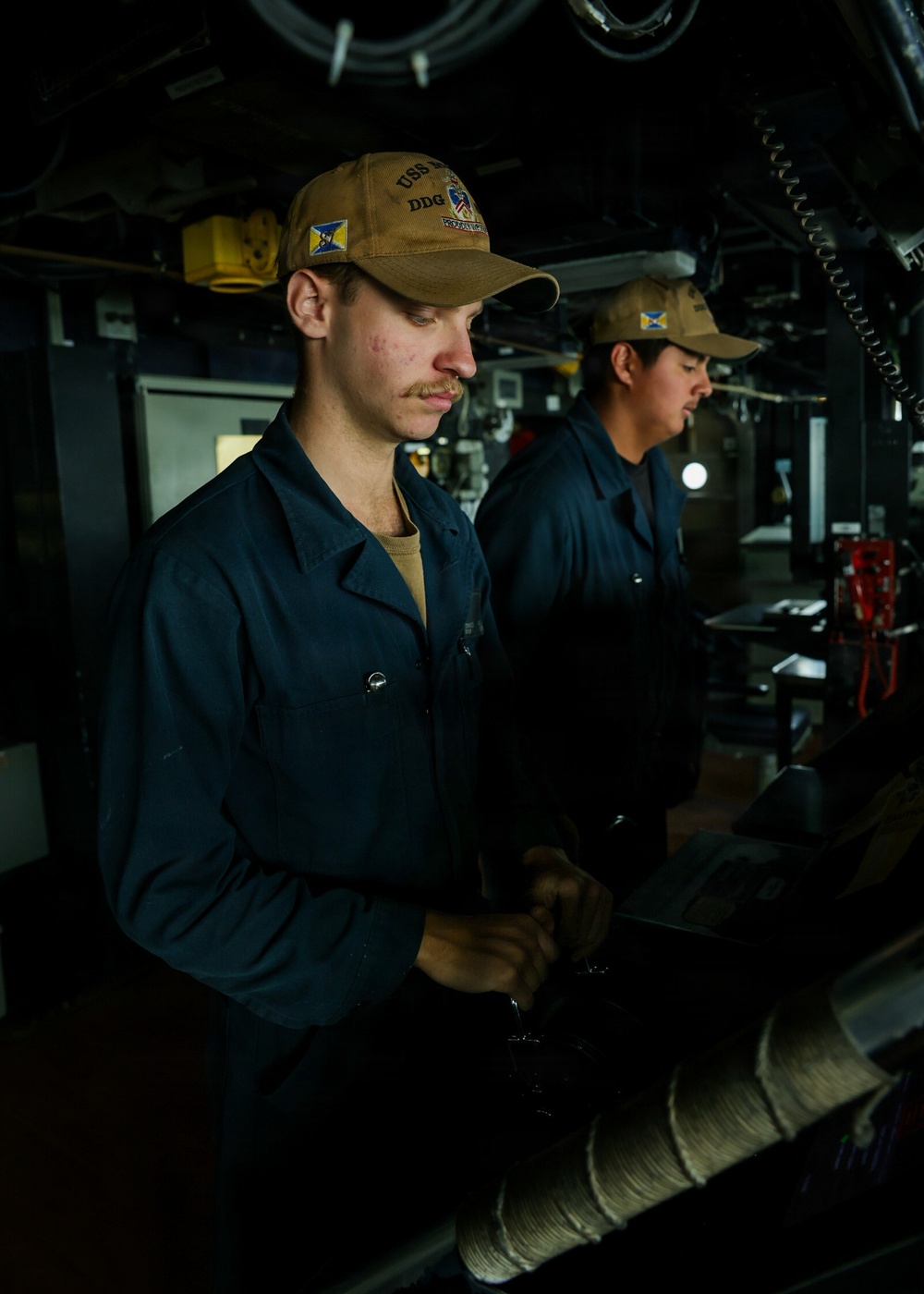Two naval personnel are stationed at what appears to be the bridge or helm of a U.S. Navy destroyer (DDG). Both are wearing matching tan/coyote brown ball caps bearing 'USS' and 'DDG' designations, along with dark navy blue coveralls. They appear to be on watch duty, manning control stations aboard the vessel.