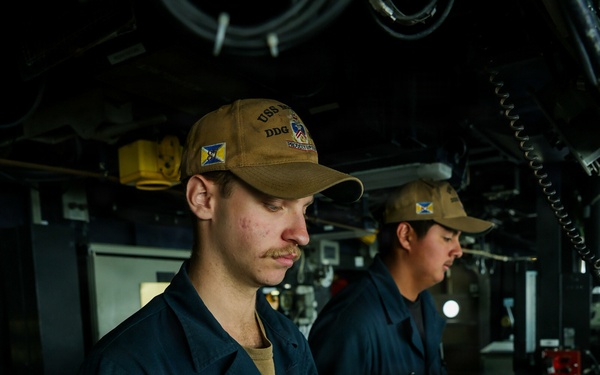 Two naval personnel are stationed at what appears to be the bridge or helm of a U.S. Navy destroyer (DDG). Both are wearing matching tan/coyote brown ball caps bearing 'USS' and 'DDG' designations, along with dark navy blue coveralls. They appear to be on watch duty, manning control stations aboard the vessel.