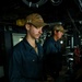 Two naval personnel are stationed at what appears to be the bridge or helm of a U.S. Navy destroyer (DDG). Both are wearing matching tan/coyote brown ball caps bearing 'USS' and 'DDG' designations, along with dark navy blue coveralls. They appear to be on watch duty, manning control stations aboard the vessel.