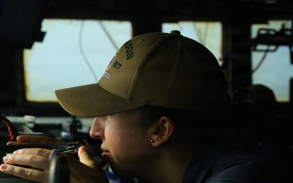 A person wearing a olive/tan baseball cap and dark jacket is looking through a navigational instrument, likely a gyrocompass repeater or bearing circle, aboard what appears to be a naval vessel's bridge. The individual is wearing a smartwatch and a small earring, and is focused on taking a bearing or sighting reading. The setting is clearly a ship's bridge with windows overlooking open water and various electronic equipment visible in the background.