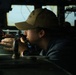 A person wearing a olive/tan baseball cap and dark jacket is looking through a navigational instrument, likely a gyrocompass repeater or bearing circle, aboard what appears to be a naval vessel's bridge. The individual is wearing a smartwatch and a small earring, and is focused on taking a bearing or sighting reading. The setting is clearly a ship's bridge with windows overlooking open water and various electronic equipment visible in the background.