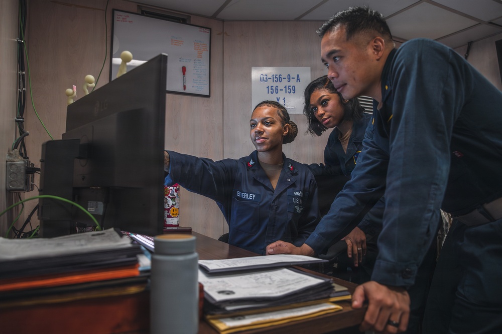 Three U.S. Navy personnel in navy blue uniforms are gathered around a computer monitor in what appears to be an onboard office or administrative workspace. The sailor in the foreground, identified by name tape as 'BEVERLEY,' is pointing at the screen while two colleagues look on. The setting suggests a working military environment aboard a naval vessel.