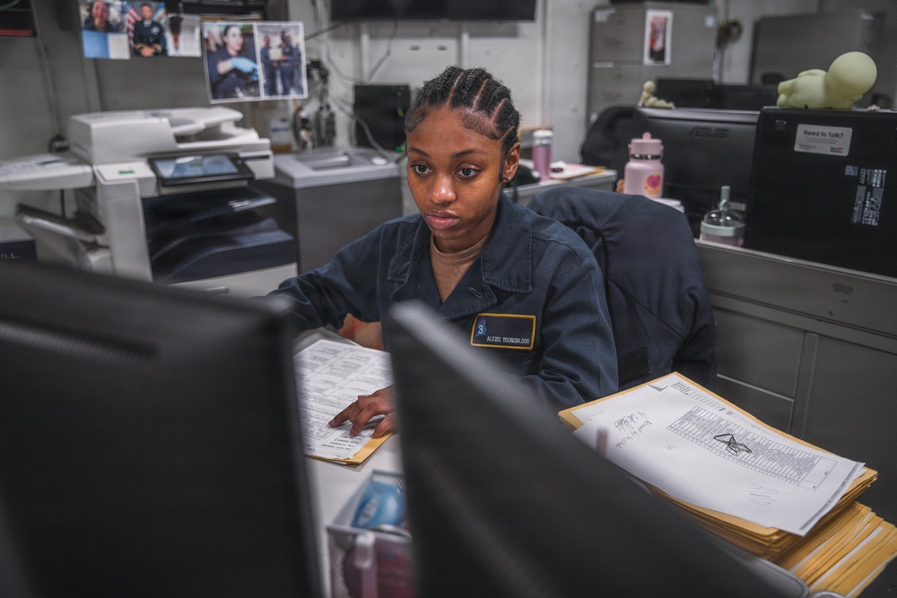 A U.S. Navy service member named Alexis Youngblood, wearing a dark blue Navy working uniform, is seated at a desk working at a computer workstation. The office environment appears to be aboard a naval vessel or military installation, with administrative paperwork, a multifunction printer, and personal photos visible in the background. The scene depicts routine administrative or clerical duties being performed.