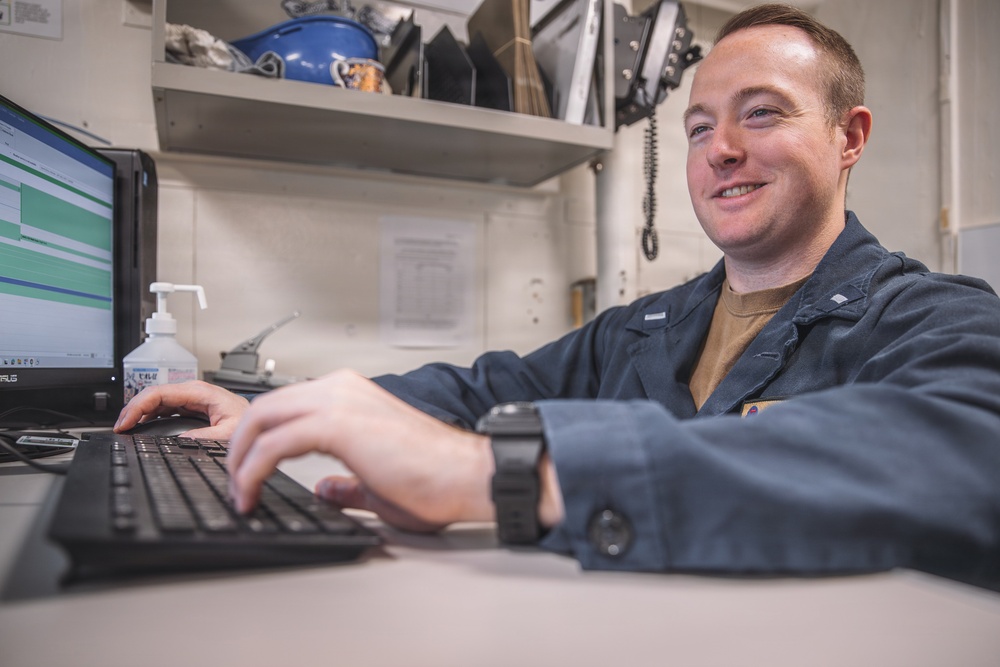 A young man wearing dark blue coveralls is seated at a workstation, smiling while typing on a keyboard. The setting appears to be a military or industrial workspace, suggested by the coverall uniform and utilitarian environment. A computer monitor displaying a green-highlighted interface is visible in front of him.