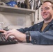 A young man wearing dark blue coveralls is seated at a workstation, smiling while typing on a keyboard. The setting appears to be a military or industrial workspace, suggested by the coverall uniform and utilitarian environment. A computer monitor displaying a green-highlighted interface is visible in front of him.