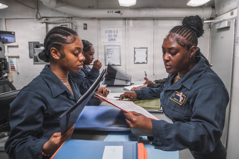 Three individuals in dark blue military/naval uniforms are working in what appears to be an administrative office aboard a naval vessel. Two personnel in the foreground are reviewing documents together across a counter, while a third works independently in the background. The setting shows a shipboard administrative workspace with paperwork, binders, and office equipment.