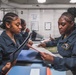Three individuals in dark blue military/naval uniforms are working in what appears to be an administrative office aboard a naval vessel. Two personnel in the foreground are reviewing documents together across a counter, while a third works independently in the background. The setting shows a shipboard administrative workspace with paperwork, binders, and office equipment.