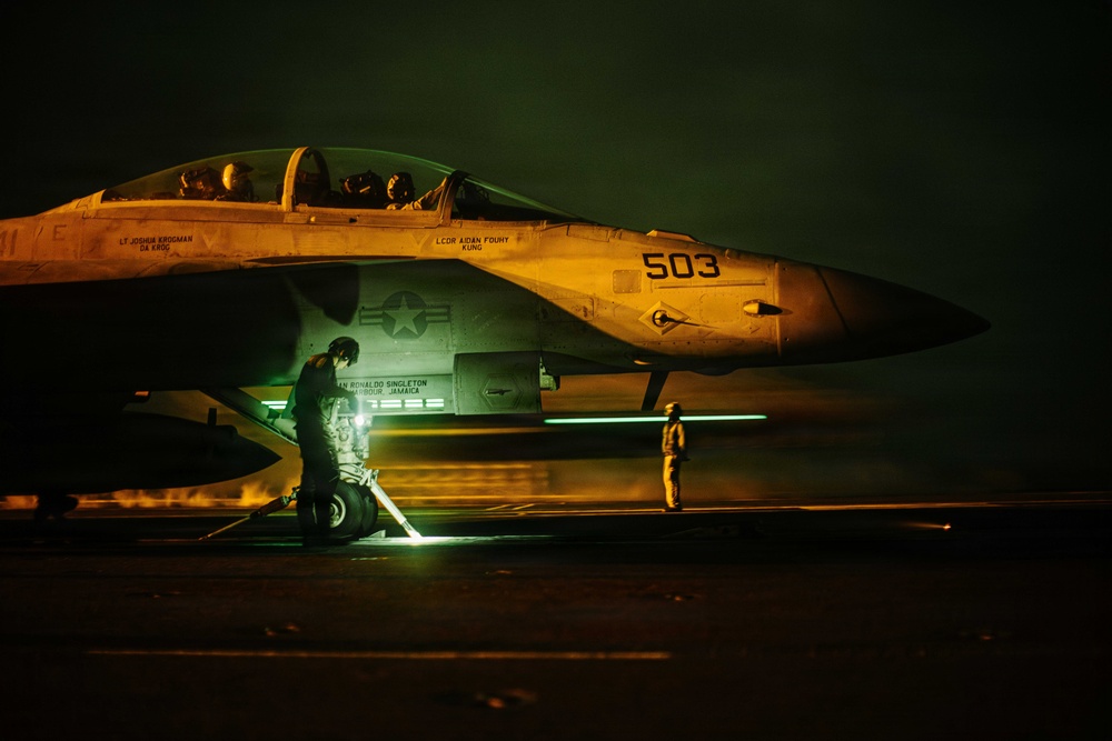 A U.S. Navy F/A-18 Super Hornet (bureau number 503) is photographed on what appears to be an aircraft carrier flight deck during nighttime operations. Ground crew personnel are conducting pre-flight or post-flight checks under dramatic orange and green lighting. Two crew members are visible in the cockpit, and at least two deck crew members are working around the aircraft.