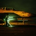 A U.S. Navy F/A-18 Super Hornet (bureau number 503) is photographed on what appears to be an aircraft carrier flight deck during nighttime operations. Ground crew personnel are conducting pre-flight or post-flight checks under dramatic orange and green lighting. Two crew members are visible in the cockpit, and at least two deck crew members are working around the aircraft.