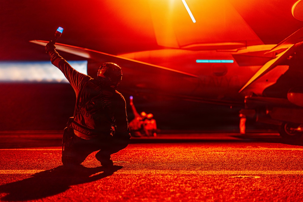A military flight deck crew member is kneeling on a flight deck at night, raising a blue illuminated wand/signal device toward a military aircraft. The scene is bathed in intense red/orange lighting, characteristic of night operations on an aircraft carrier flight deck. Additional crew members are visible in the background near the aircraft.