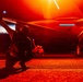 A military flight deck crew member is kneeling on a flight deck at night, raising a blue illuminated wand/signal device toward a military aircraft. The scene is bathed in intense red/orange lighting, characteristic of night operations on an aircraft carrier flight deck. Additional crew members are visible in the background near the aircraft.
