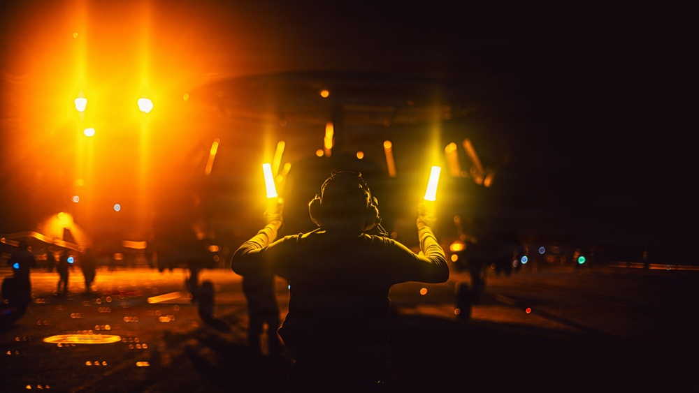 A person is seen from behind holding two illuminated orange/yellow light wands in an outstretched position, consistent with aircraft marshalling or flight deck operations at night. The scene appears to take place on what looks like a military aircraft carrier flight deck, with bright orange lighting, aircraft, and other personnel visible in the background. The dramatic lighting and dark environment suggest nighttime flight operations.