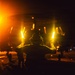 A person is seen from behind holding two illuminated orange/yellow light wands in an outstretched position, consistent with aircraft marshalling or flight deck operations at night. The scene appears to take place on what looks like a military aircraft carrier flight deck, with bright orange lighting, aircraft, and other personnel visible in the background. The dramatic lighting and dark environment suggest nighttime flight operations.