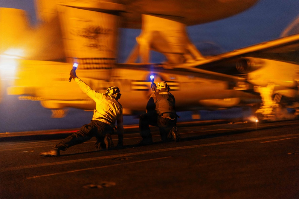 Two naval flight deck crew members are directing an aircraft during night operations on what appears to be an aircraft carrier flight deck. Both personnel are holding illuminated blue wands/flashlights used for signaling, with one crew member in a characteristic low crouching launch signal pose. The image is motion-blurred, capturing the dynamic and fast-paced nature of carrier flight deck operations at dusk or nighttime.