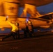 Two naval flight deck crew members are directing an aircraft during night operations on what appears to be an aircraft carrier flight deck. Both personnel are holding illuminated blue wands/flashlights used for signaling, with one crew member in a characteristic low crouching launch signal pose. The image is motion-blurred, capturing the dynamic and fast-paced nature of carrier flight deck operations at dusk or nighttime.