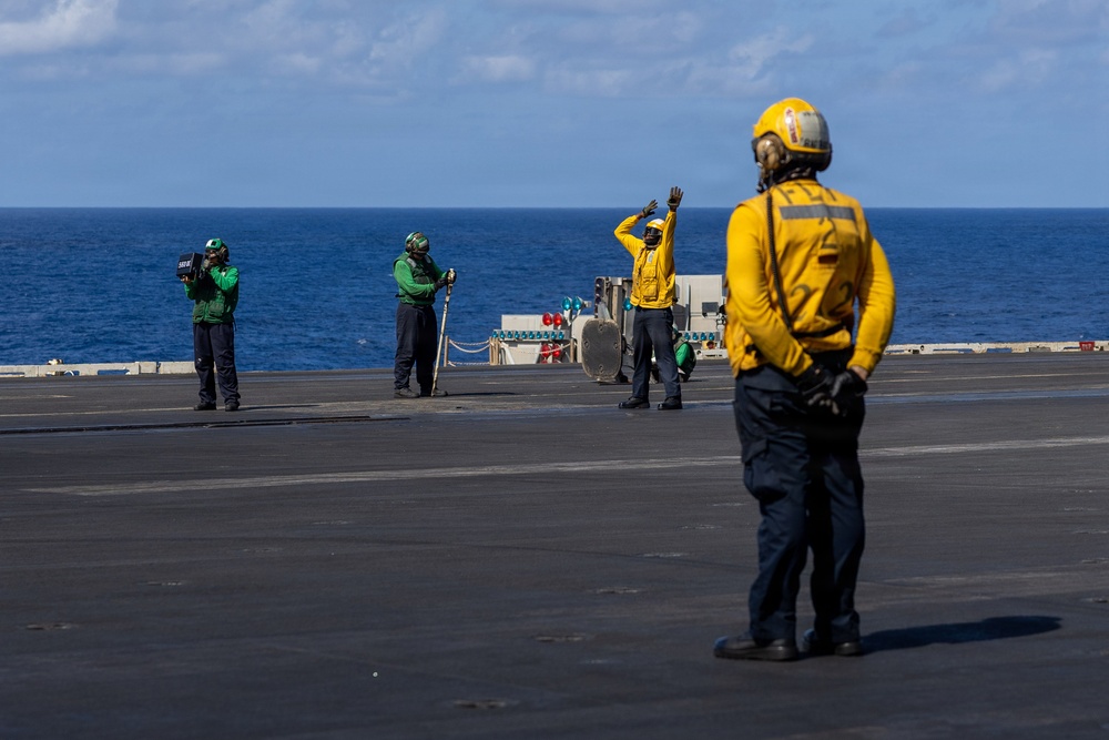 The image shows flight deck crew members aboard what appears to be a U.S. Navy aircraft carrier operating at sea. Personnel are wearing color-coded jerseys and helmets indicating their specific roles in flight deck operations. The scene depicts standard carrier flight deck procedures with crew members in communication and signaling positions.