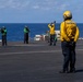 The image shows flight deck crew members aboard what appears to be a U.S. Navy aircraft carrier operating at sea. Personnel are wearing color-coded jerseys and helmets indicating their specific roles in flight deck operations. The scene depicts standard carrier flight deck procedures with crew members in communication and signaling positions.
