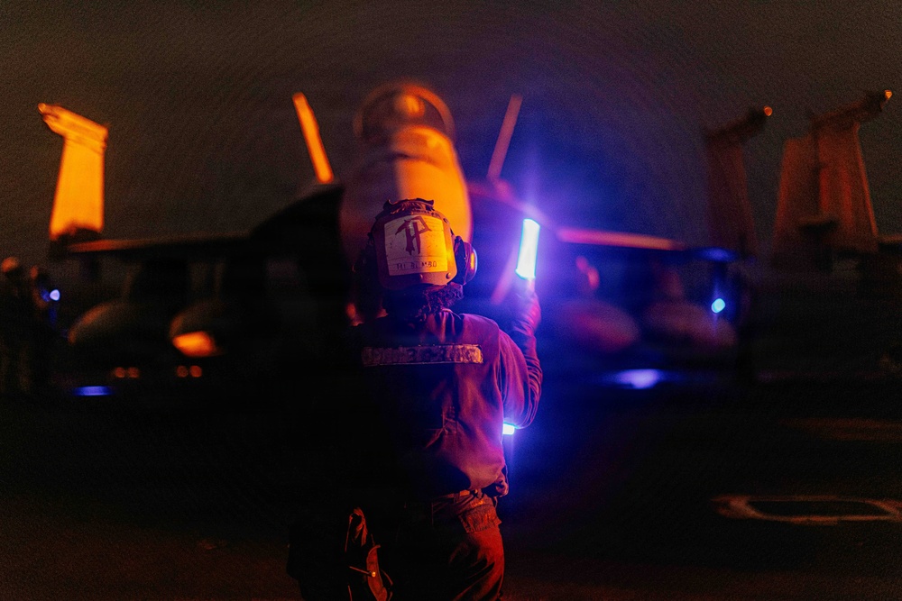A naval flight deck crew member is directing aircraft during nighttime flight operations, likely aboard an aircraft carrier. The individual is holding an illuminated wand and facing a military jet aircraft, identifiable as an F/A-18 Hornet or Super Hornet by its twin-tail configuration. The scene is dramatically lit with orange, blue, and white light sources against a dark background.