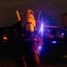 A naval flight deck crew member is directing aircraft during nighttime flight operations, likely aboard an aircraft carrier. The individual is holding an illuminated wand and facing a military jet aircraft, identifiable as an F/A-18 Hornet or Super Hornet by its twin-tail configuration. The scene is dramatically lit with orange, blue, and white light sources against a dark background.