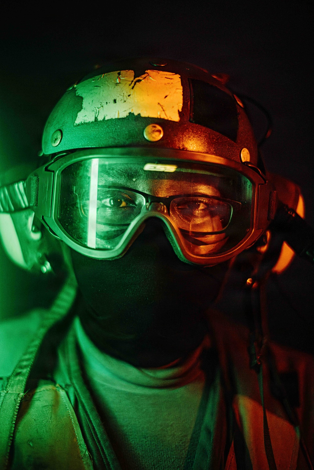 A close-up portrait of a person wearing tactical military-style gear, illuminated by contrasting green and orange/red colored lighting. The subject is equipped with a ballistic helmet, protective goggles, and a balaclava face covering. The dramatic dual-tone lighting creates a striking visual effect against a dark background.