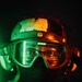 A close-up portrait of a person wearing tactical military-style gear, illuminated by contrasting green and orange/red colored lighting. The subject is equipped with a ballistic helmet, protective goggles, and a balaclava face covering. The dramatic dual-tone lighting creates a striking visual effect against a dark background.