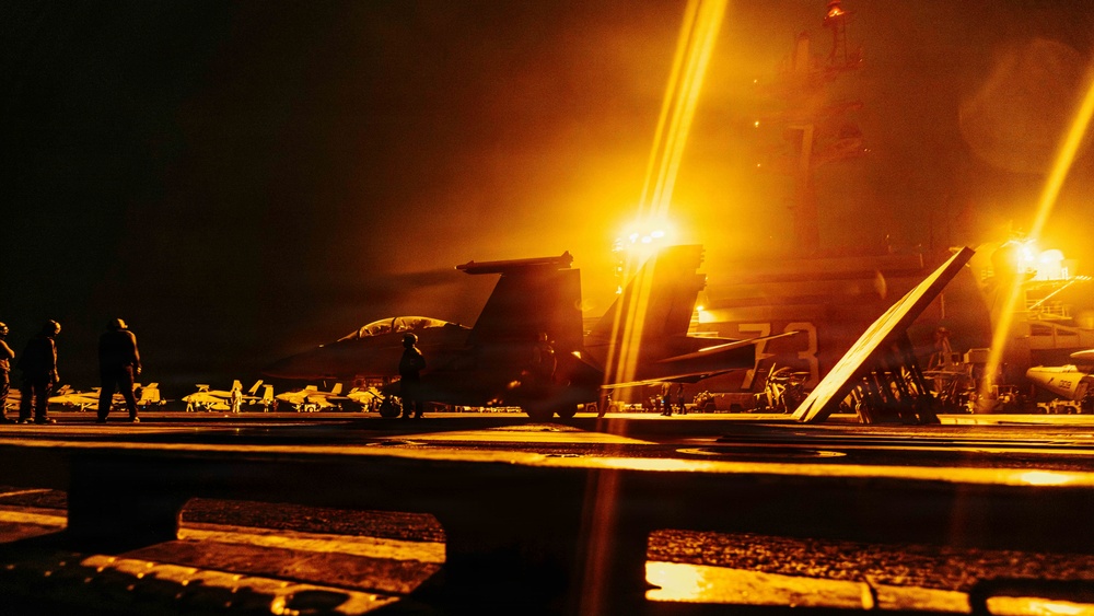 A nighttime scene on what appears to be a naval aircraft carrier flight deck, illuminated by intense orange-yellow artificial lighting. Military jet aircraft are visible on the deck alongside several silhouetted personnel. The dramatic lighting and low-angle perspective create a high-contrast, atmospheric composition.