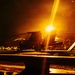A nighttime scene on what appears to be a naval aircraft carrier flight deck, illuminated by intense orange-yellow artificial lighting. Military jet aircraft are visible on the deck alongside several silhouetted personnel. The dramatic lighting and low-angle perspective create a high-contrast, atmospheric composition.
