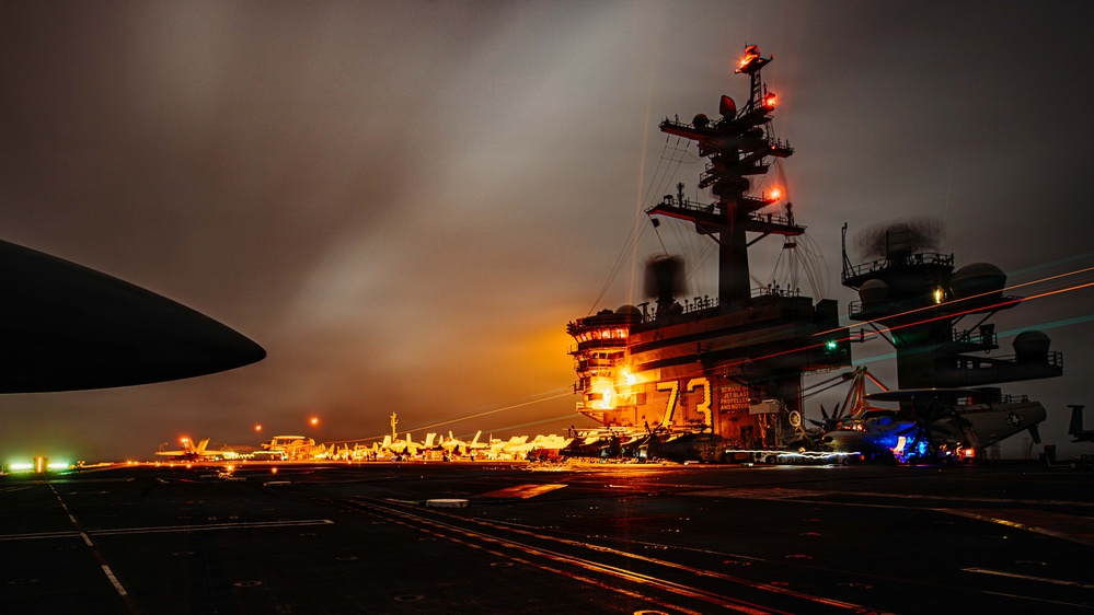 A nighttime photograph taken on the flight deck of USS George Washington (CVN-73), a Nimitz-class aircraft carrier. The image captures the island superstructure illuminated with orange lighting, with the hull number '73' clearly visible. Multiple aircraft are parked on the flight deck, and light trails from moving vehicles or personnel are visible, suggesting a long-exposure photograph.