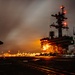 A nighttime photograph taken on the flight deck of USS George Washington (CVN-73), a Nimitz-class aircraft carrier. The image captures the island superstructure illuminated with orange lighting, with the hull number '73' clearly visible. Multiple aircraft are parked on the flight deck, and light trails from moving vehicles or personnel are visible, suggesting a long-exposure photograph.