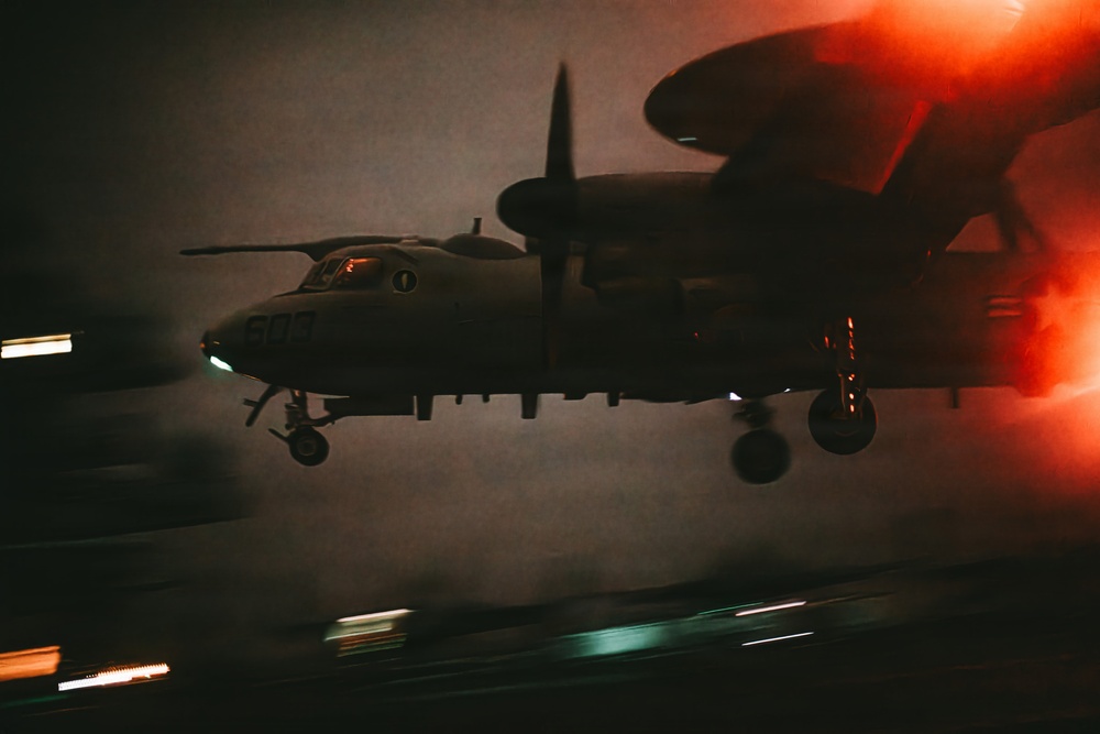 A military turboprop aircraft, identifiable as a Grumman E-2 Hawkeye based on its distinctive rotodome radar disc above the fuselage, is captured during a nighttime carrier landing or low-altitude approach. The image is dramatically lit with red ambient light and motion blur from runway or deck lighting below, suggesting a carrier deck environment. The aircraft's landing gear is deployed and navigation lights are visible.
