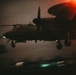 A military turboprop aircraft, identifiable as a Grumman E-2 Hawkeye based on its distinctive rotodome radar disc above the fuselage, is captured during a nighttime carrier landing or low-altitude approach. The image is dramatically lit with red ambient light and motion blur from runway or deck lighting below, suggesting a carrier deck environment. The aircraft's landing gear is deployed and navigation lights are visible.
