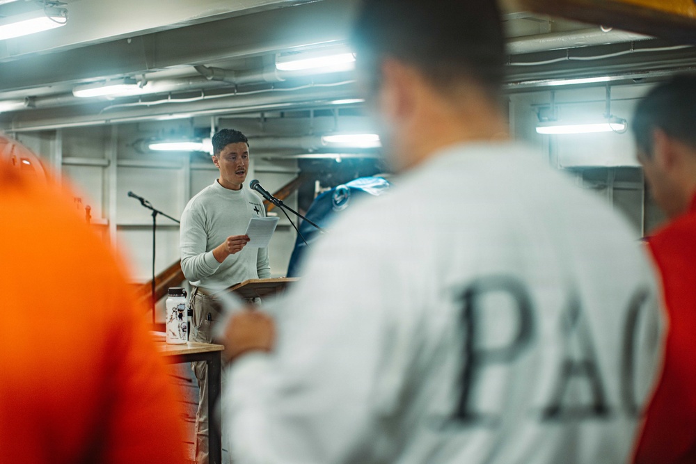 A person stands at a podium or lectern holding papers and speaking into a microphone in what appears to be an industrial or ship interior space. An audience is visible in the foreground, with one person wearing a sweatshirt with 'PAC' visible on the back. The setting has fluorescent lighting and metal walls consistent with a vessel or warehouse environment.