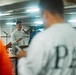 A person stands at a podium or lectern holding papers and speaking into a microphone in what appears to be an industrial or ship interior space. An audience is visible in the foreground, with one person wearing a sweatshirt with 'PAC' visible on the back. The setting has fluorescent lighting and metal walls consistent with a vessel or warehouse environment.
