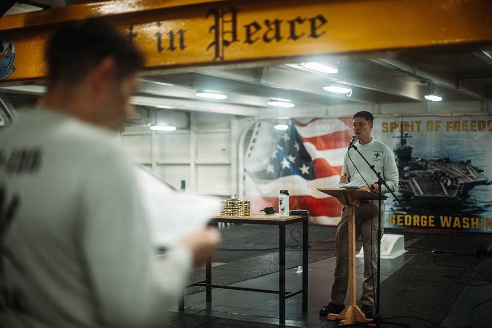 A person stands at a podium with a microphone aboard what appears to be the USS George Washington aircraft carrier, likely conducting a religious service or memorial ceremony. A second person in the foreground holds papers, possibly reading or participating in the service. The setting features an American flag mural, the ship's name 'George Washington,' and a beam with the partial text 'in Peace' visible overhead.