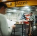 A person stands at a podium with a microphone aboard what appears to be the USS George Washington aircraft carrier, likely conducting a religious service or memorial ceremony. A second person in the foreground holds papers, possibly reading or participating in the service. The setting features an American flag mural, the ship's name 'George Washington,' and a beam with the partial text 'in Peace' visible overhead.
