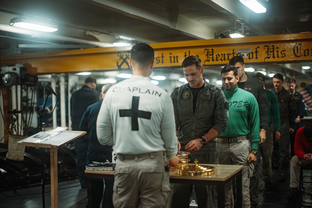 A military chaplain is conducting a religious service, likely a communion ceremony, aboard a naval vessel. The chaplain, wearing a white shirt with 'CHAPLAIN' and a black cross on the back, stands at a small table with gold communion vessels. Several service members in various uniforms are gathered and participating in the service.