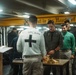 A military chaplain is conducting a religious service, likely a communion ceremony, aboard a naval vessel. The chaplain, wearing a white shirt with 'CHAPLAIN' and a black cross on the back, stands at a small table with gold communion vessels. Several service members in various uniforms are gathered and participating in the service.