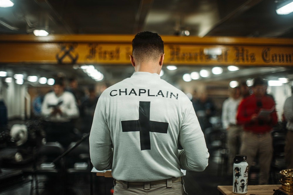 A military or institutional chaplain is seen from behind, wearing a white long-sleeve shirt with 'CHAPLAIN' printed on the back along with a black cross symbol. The setting appears to be an indoor gathering space, possibly aboard a naval vessel or military facility, with a yellow overhead beam bearing a partially visible inscription. A group of people are assembled in the background, suggesting a religious service or ceremony is taking place.