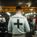 A military or institutional chaplain is seen from behind, wearing a white long-sleeve shirt with 'CHAPLAIN' printed on the back along with a black cross symbol. The setting appears to be an indoor gathering space, possibly aboard a naval vessel or military facility, with a yellow overhead beam bearing a partially visible inscription. A group of people are assembled in the background, suggesting a religious service or ceremony is taking place.