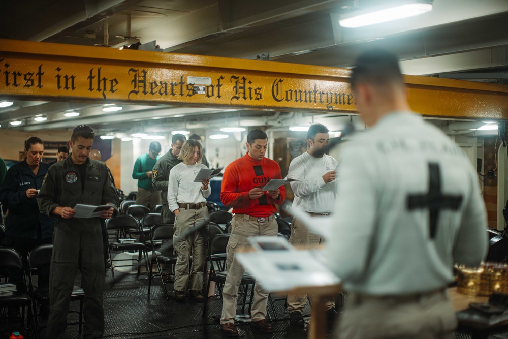 A religious service is being conducted aboard a U.S. Navy vessel, with a chaplain (identifiable by the cross on their back) leading a group of sailors in what appears to be a worship or prayer service. The participants are standing and reading from printed materials, likely programs or hymnals. A yellow beam overhead bears the inscription 'First in the Hearts of His Countrymen,' a phrase associated with George Washington.