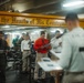A religious service is being conducted aboard a U.S. Navy vessel, with a chaplain (identifiable by the cross on their back) leading a group of sailors in what appears to be a worship or prayer service. The participants are standing and reading from printed materials, likely programs or hymnals. A yellow beam overhead bears the inscription 'First in the Hearts of His Countrymen,' a phrase associated with George Washington.