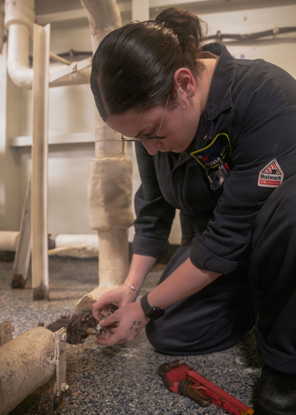 A person wearing a dark blue Bulwark FR (flame-resistant) coverall is kneeling on the floor working on a pipe fitting or plumbing connection at floor level. A name tag reading 'OLIVIA LOF...' is visible on the chest. A red pipe wrench lies nearby on the floor, and insulated piping is visible in the background.