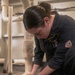 A person wearing a dark blue Bulwark FR (flame-resistant) coverall is kneeling on the floor working on a pipe fitting or plumbing connection at floor level. A name tag reading 'OLIVIA LOF...' is visible on the chest. A red pipe wrench lies nearby on the floor, and insulated piping is visible in the background.